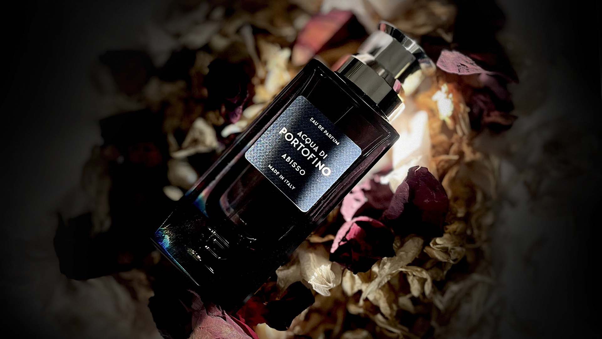 Dark glass Acqua di Portofino Abisso Eau de Parfum bottle with silver cap resting among dried red rose petals and botanical textures under dramatic spotlighting.