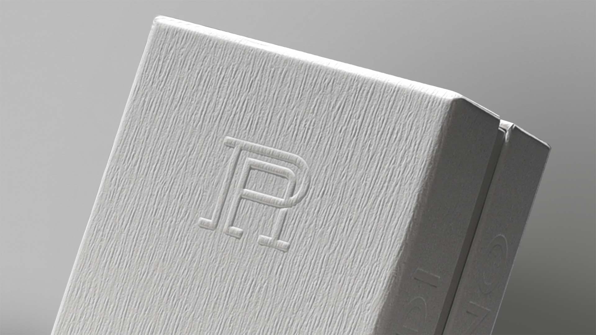 Macro closeup of paper grain texture and embossed monogram detailing on rigid secondary box packaging.