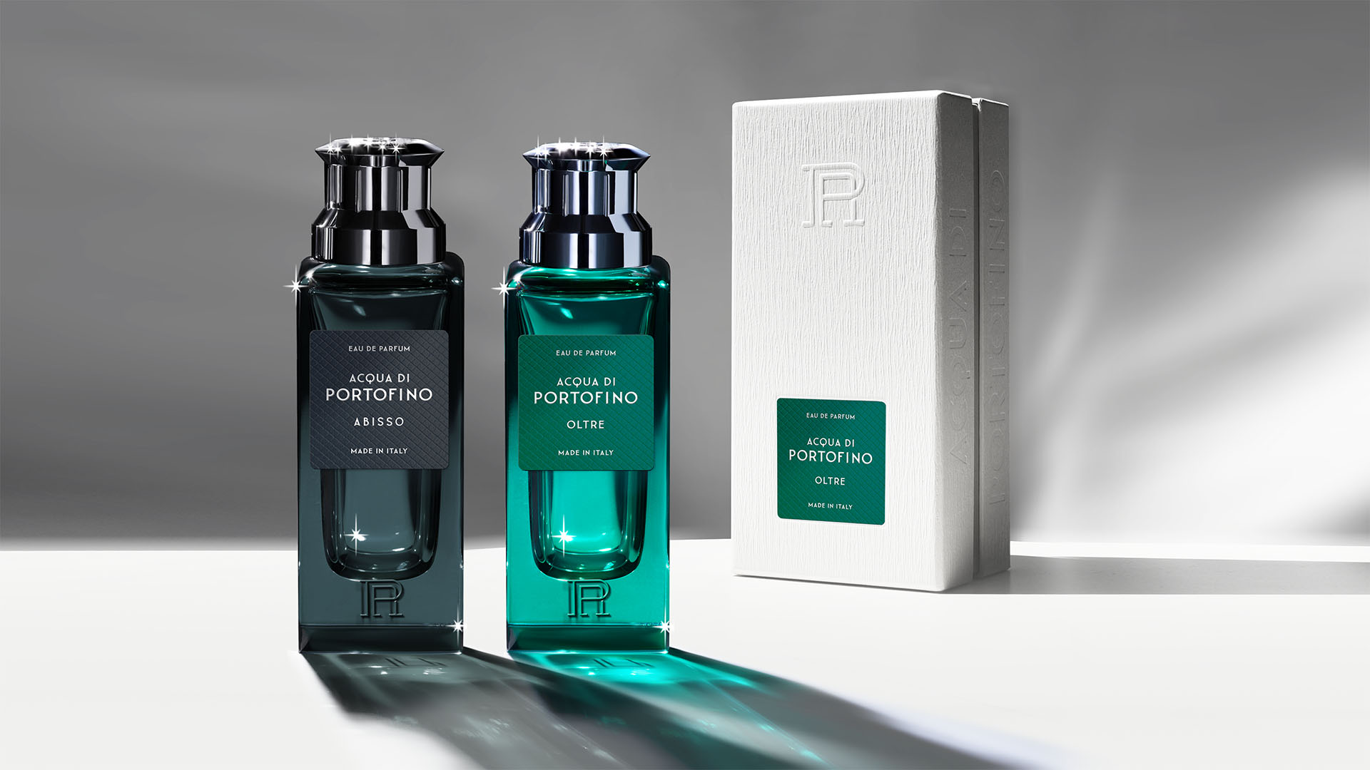 Abisso and Oltre fragrance bottles in dark teal glass standing alongside embossed white outer packaging.